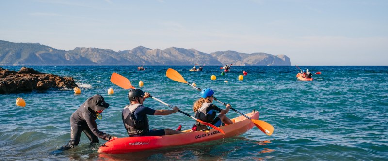 People kayaking in the ocean near a rocky shore with distant mountains.