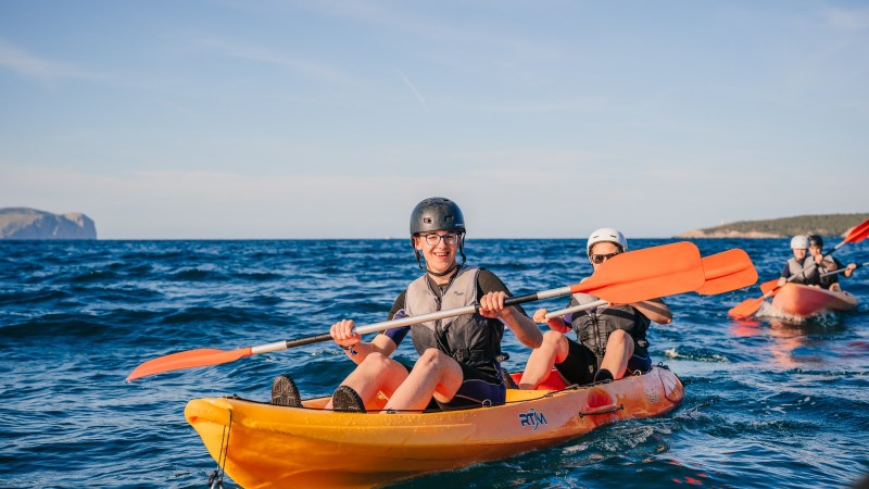 Group of people kayaking on open water, wearing helmets and life vests, with a clear sky.