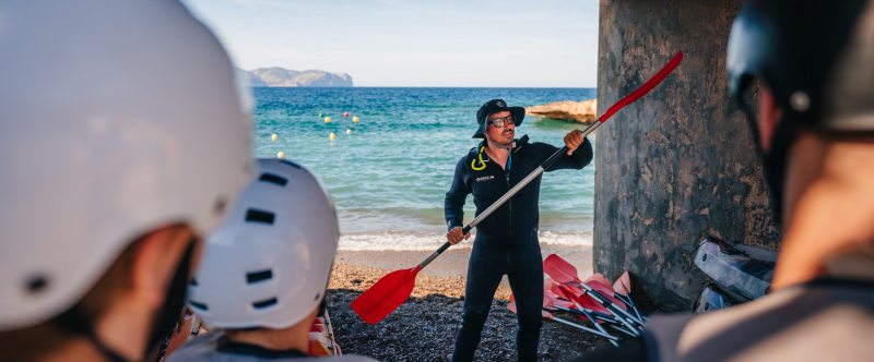 Instructor in wetsuit teaching kayaking by the sea to helmeted group.