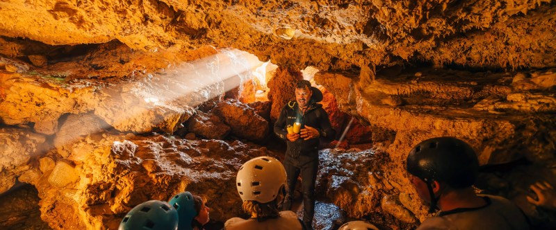 Group of people with helmets in a cave, illuminated by sunlight.