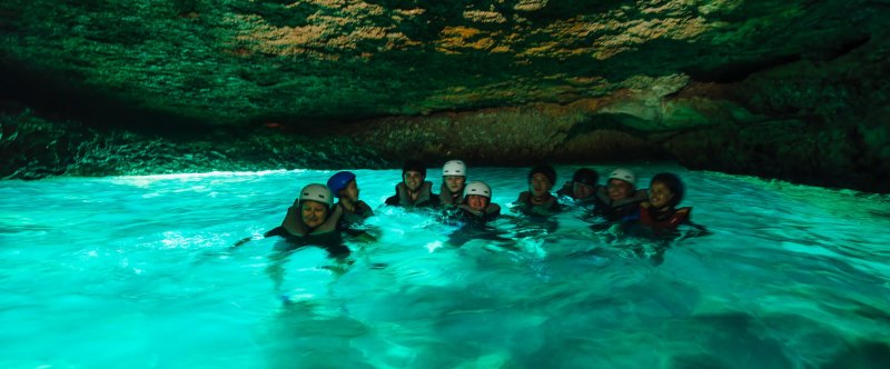 Group wearing helmets swimming in a turquoise-lit cave.