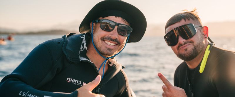 Two men in wetsuits and sunglasses smiling and gesturing, sitting by the sea.