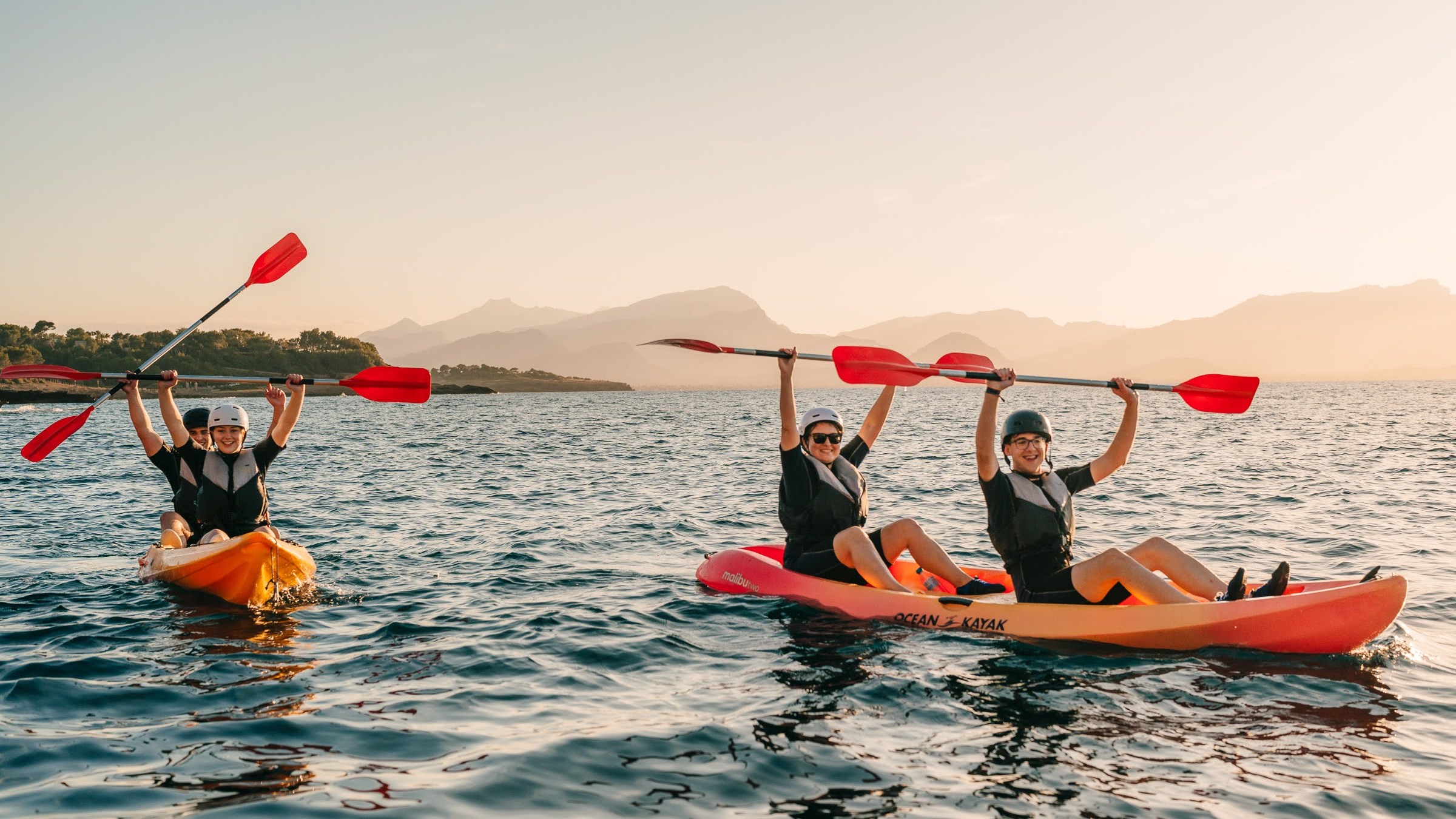 Four people kayaking on a lake at sunset, raising paddles in the air, with mountains in the background.