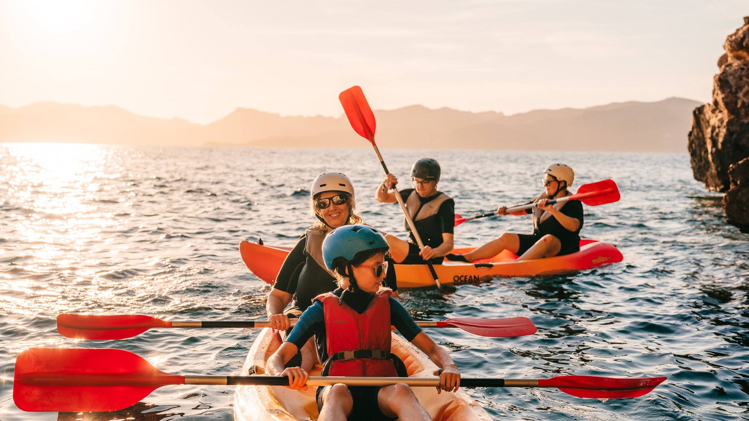 Group of kayakers paddling on the ocean near a rocky shore under a clear sky at sunset.