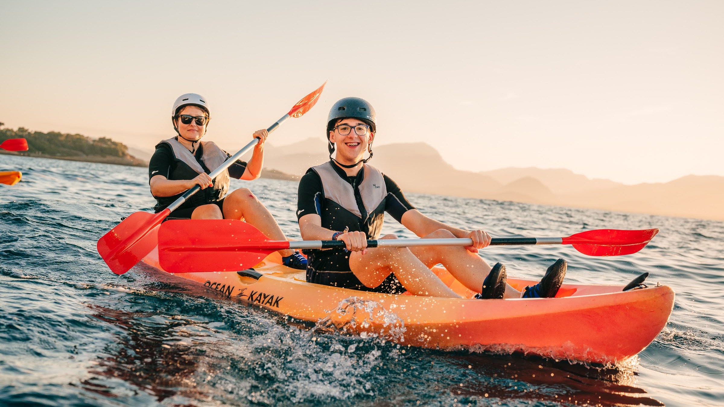 Two people kayaking in the ocean at sunset, wearing helmets and life vests.
