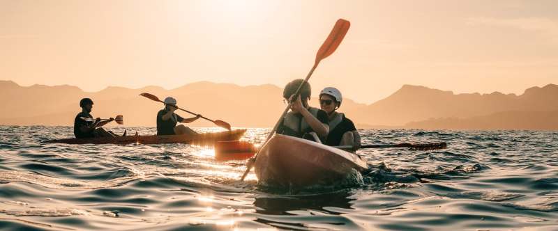 People kayaking on a calm sea at sunset with mountains in the background.