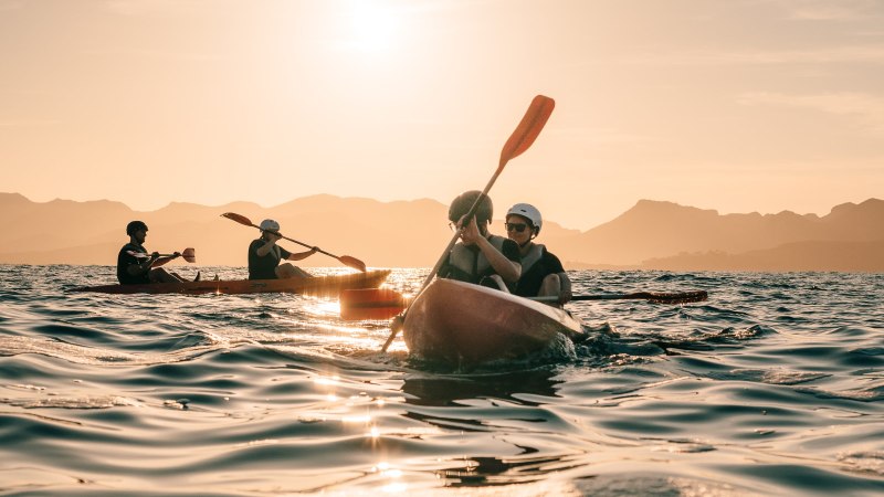 People kayaking on a calm sea at sunset with mountains in the background.