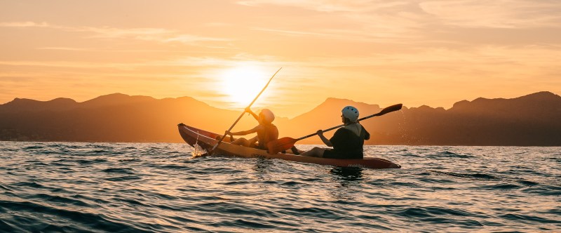 Two people kayaking on the ocean at sunset with mountains in the background.