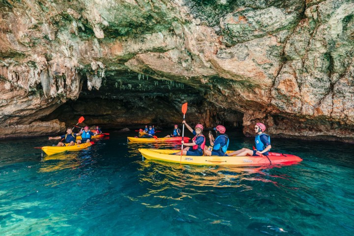 People kayaking in a large cave with rocky ceiling and turquoise water.
