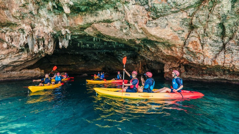 People kayaking in a large cave with rocky ceiling and turquoise water.