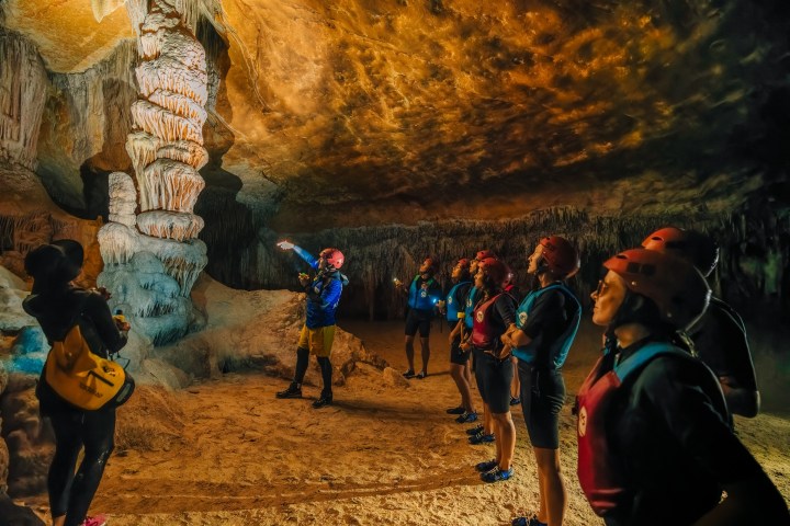 Group of people with helmets and vests exploring a cave with stalactites.