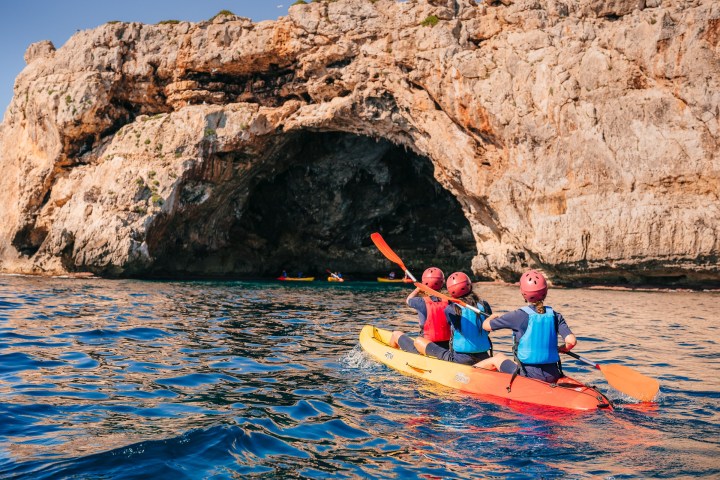 Three people kayaking toward a large rock cave on a sunny day.